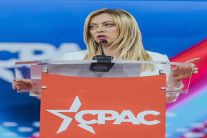 Eine Frau in einem weißen Kleid steht an einem Rednerpult mit einem Mikrofon, unscharfer Hintergrund legt den Fokus auf sie, wahrscheinlich eine Politikwissenschaftlerin.