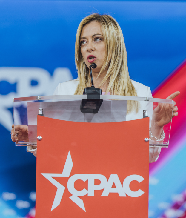 Eine Frau in einem weißen Kleid steht an einem Rednerpult mit einem Mikrofon, unscharfer Hintergrund legt den Fokus auf sie, wahrscheinlich eine Politikwissenschaftlerin.