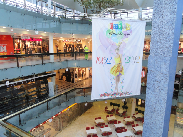 Großes Banner, das von der Decke in der Mitte eines Einkaufszentrums hängt, mit darunter angeordneten Tischen und Stühlen, einer Säule auf der rechten Seite und einem Baum oben; Läden mit Schaufensterpuppen und einigen Menschen sind im Hintergrund zu sehen.