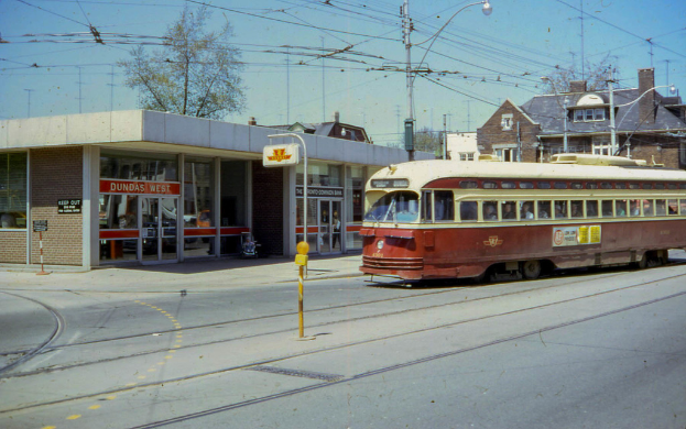 Ein Zug ist auf der Straße zu sehen, mit Gebäuden und Bäumen im Hintergrund, Oberleitungen und Masten sowie einem Himmel darüber.