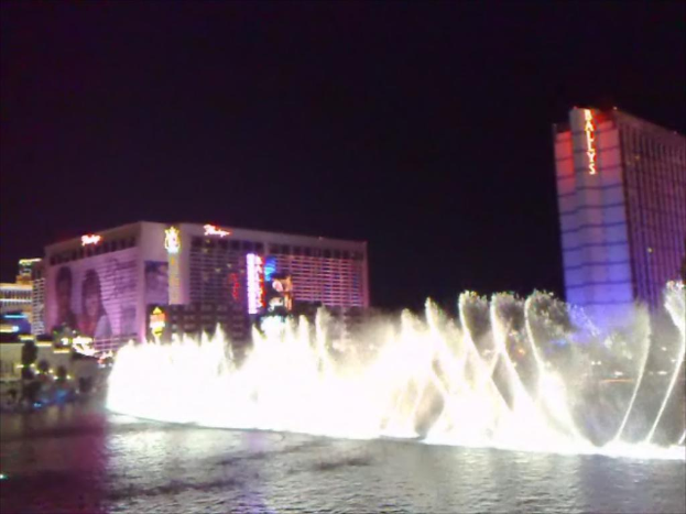 Ein Brunnen mit Wasser am Boden, umgeben von Gebäuden, Plakaten, Bildschirmen, Lichtern, Pflanzen und Himmel im Hintergrund.