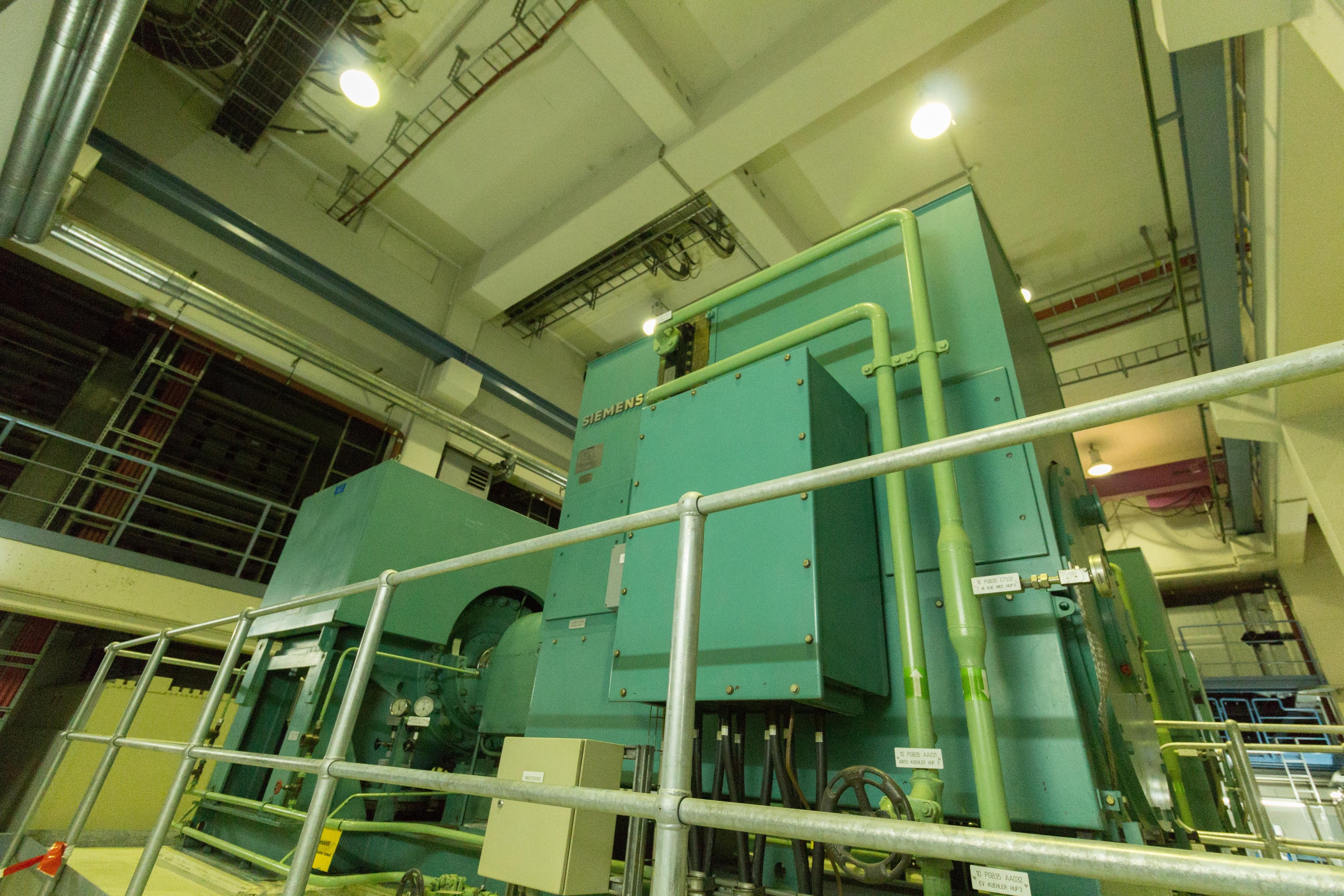 Großer industrieller Wärmetauscher in einem Gebäude, umgeben von Geländern, Rohren, Ventilen und Beleuchtung.