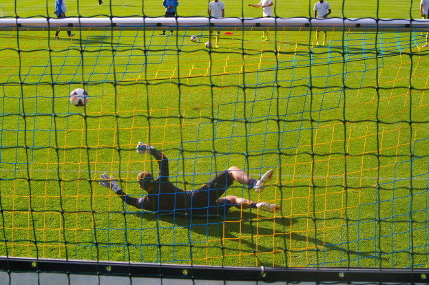 Ein Fußballspieler hält vor einem Tor, mit einem Netz im Vordergrund und einigen Menschen im Hintergrund, während mehrere Fußballs auf dem Boden verstreut sind.