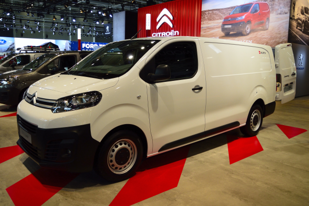 Ein weißer Citroen Berlingo Van wird auf einer Autauschau präsentiert, umgeben von schwarzen Schlafzimmermöbeln, während die Wände mit Brettern und Lichtern geschmückt sind.
