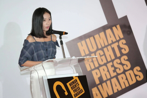Eine Frau in einem schwarzen Kleid steht an einem Podium mit einem Mikrofon und hält eine Rede vor einem Banner "Human Rights Press Awards", ihr Haar ist zurückgebunden und ihr Gesichtsausdruck ernst und konzentriert.