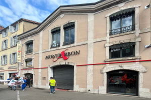 Eine Gruppe von Menschen mit Fahnen geht eine Straße entlang vor einem Gebäude mit einer Schilderwand, die den Text 'Montbron' trägt, einem Straßenschild und einer Fahne mit Bildern und Text unter einem bewölkten Himmel.