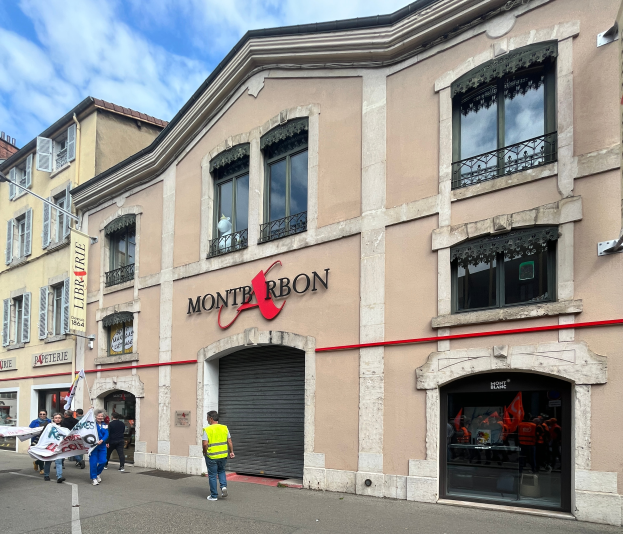 Eine Gruppe von Menschen mit Fahnen geht eine Straße entlang vor einem Gebäude mit einer Schilderwand, die den Text 'Montbron' trägt, einem Straßenschild und einer Fahne mit Bildern und Text unter einem bewölkten Himmel.