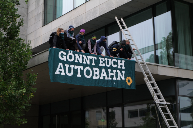 Eine Gruppe von Menschen auf dem Dach eines Gebäudes stehend, die eine Fahne halten, mit einem Baum links und Glasfenstern im Hintergrund, wahrscheinlich protestierend für die deutsche Regierung.