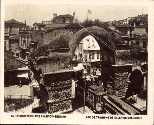 Schwarz-weiß-Foto einer Stadtstraße mit einer Bogenbrücke in der Mitte, umgeben von Gebäuden, Bäumen, Pfählen, Drähten und Menschen, mit Text unten.