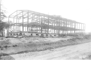 Ein Schwarz-Weiß-Foto eines im Bau befindlichen Gebäudes mit einer durchlaufenden Bahnstrecke, Bäumen auf der linken Seite und einem klaren Himmel im Hintergrund, mit Text am unteren Rand, der es als Foto des Baus einer neuen Schule identifiziert.