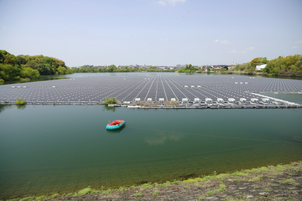 Ein kleines Boot schwimmt auf einem Gewässer, das von grünem Gras und Bäumen umgeben ist, mit Gebäuden und einem klaren blauen Himmel im Hintergrund und Solarpanelen auf der Wasseroberfläche.