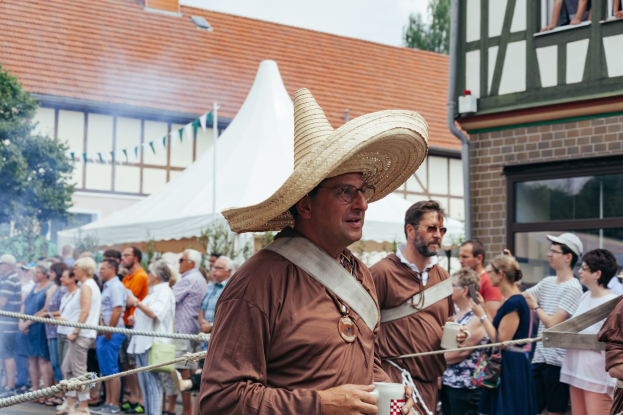 Ein Mann mit Strohhut steht vor einer Menge, hält einen Becher und hat ein Seil im Vordergrund und Gebäude, Bäume und einen klaren blauen Himmel im Hintergrund.