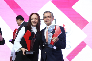 Zwei strahlende Menschen auf einer Bühne, jeder hält eine Trophäe in der Hand, mit einem Bildschirm im Hintergrund.