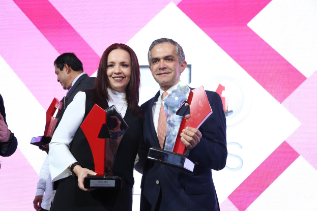 Zwei strahlende Menschen auf einer Bühne, jeder hält eine Trophäe in der Hand, mit einem Bildschirm im Hintergrund.