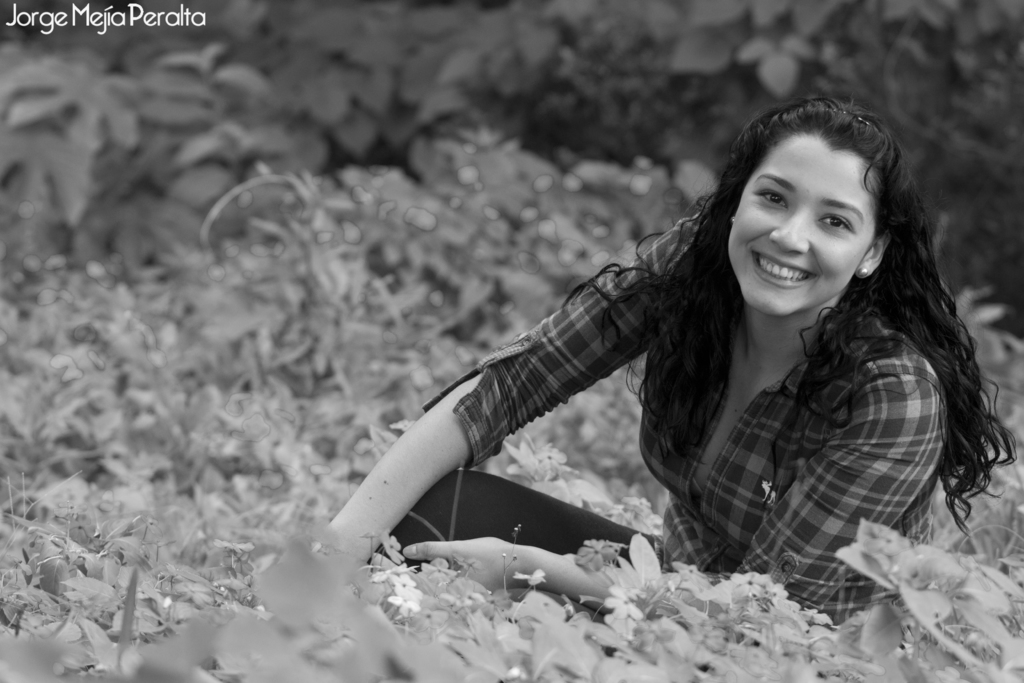 Eine Frau, die auf einem Stuhl sitzt und lächelt, mit Pflanzen im Hintergrund, Blumen unten und Text in der linken oberen Ecke.