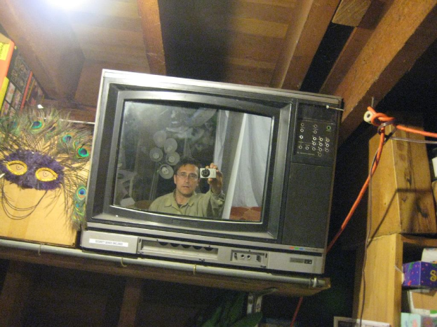 Ein Fernsehbildschirm mit einer Person umgeben von Pfauenfedern, Büchern und Kisten.