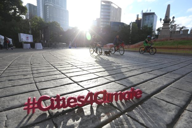 Eine Gruppe von Menschen, die auf Fahrr├Ądern eine Stra├če entlangfahren, mit der Aufschrift 'Cities4Climate' auf einem Banner, mit Geb├Ąuden, B├Ąumen, Masten, Lichtern, Bannern, Gel├Ąndern, Gras, einer Statue und Wolken im Hintergrund.