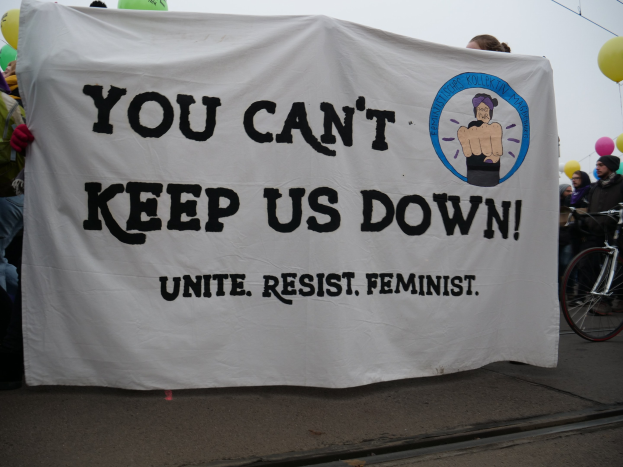 Eine Gruppe von Menschen auf einer Straße mit einem Banner, auf dem "You Can't Keep Us Down, Unite Resist Feminist" steht, mit einem Fahrrad in der Nähe und Ballons im Hintergrund unter einem klaren blauen Himmel.