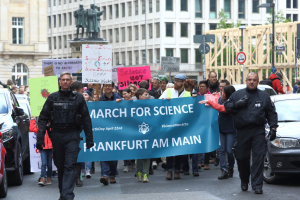 Eine Gruppe von Menschen marschiert mit einem "March for Science Frankfurt am Main"-Schild die Straße entlang, mit Autos, Gebäuden, Statuen, Laternen, Schildern und Bäumen im Hintergrund.