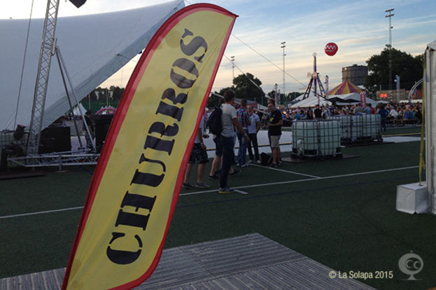 Ein buntes Volksfest-Szenario mit Zelten, einem zentralen Mast, Menschen, Bäumen, Strommasten, Himmel, Straßenbahnen auf der Straße und einem eisernen Stand auf der linken Seite.