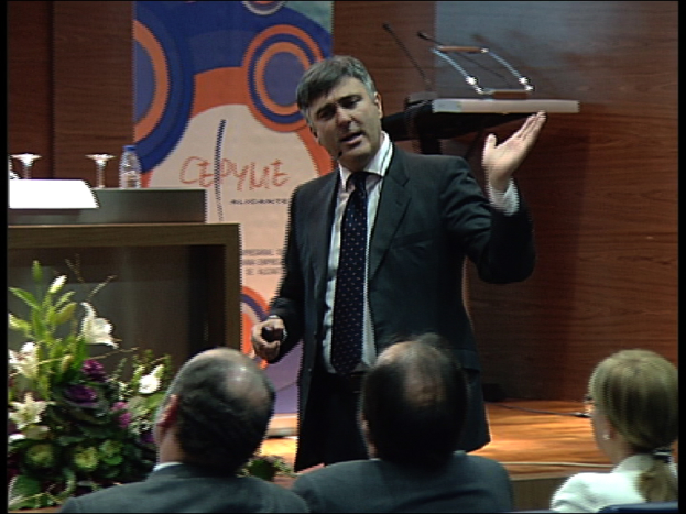 Ein Mann in einem schwarzen Blazer steht an einem Podium mit Mikrofonen, spricht in ein Handmikrofon und hat eine Blumendekoration und einen Tisch mit einem Buch und einer Wasserflasche in der Nähe, während drei Personen auf Stühlen vor ihm sitzen, vor einer braunen Wand und einer bunten Tafel mit Text.