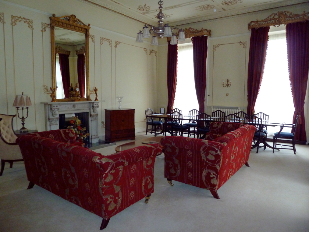 Zwei rote Sofas in einem Wohnzimmer, ein kleiner Teetisch mit einem Buch darauf, ein wandmontierter Spiegel, eine Lampe auf einem Tisch, ein Blumenbouquet vor einem Kamin, ein Esstisch mit Stühlen, von der Decke hängende Deckenleuchten, Vorhänge an einer Stange und ein leerer Stuhl.