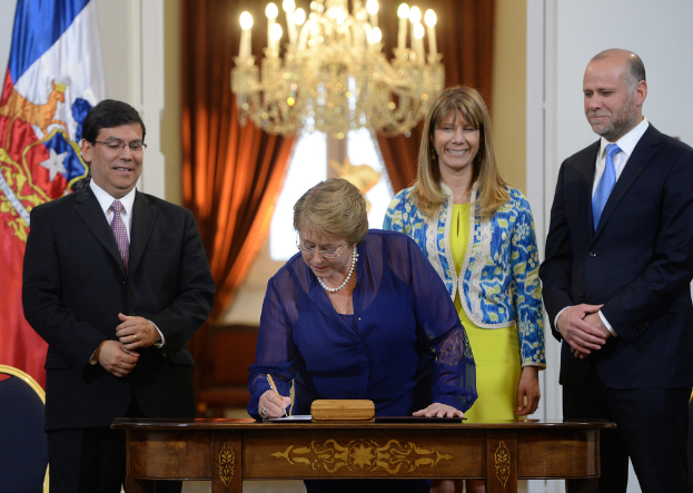 Eine Frau schreibt an einem Tisch mit Papieren und Schachteln, während hinter ihr eine Gruppe strahlender Menschen steht; an der Wand hängen eine Flagge und Vorhänge, und Kerzenlicht erhellt die Spitze des Bildes.