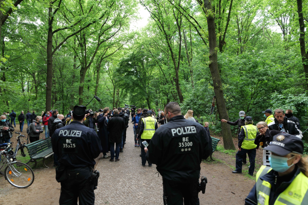 Eine Gruppe von Polizisten steht vor einer Menschenmenge, einige tragen Mützen und Masken, mit Fahrrädern und einer Bank im Vordergrund und Bäumen und Himmel im Hintergrund, wahrscheinlich bei einer Anti-Terror-Demonstration in Berlin.