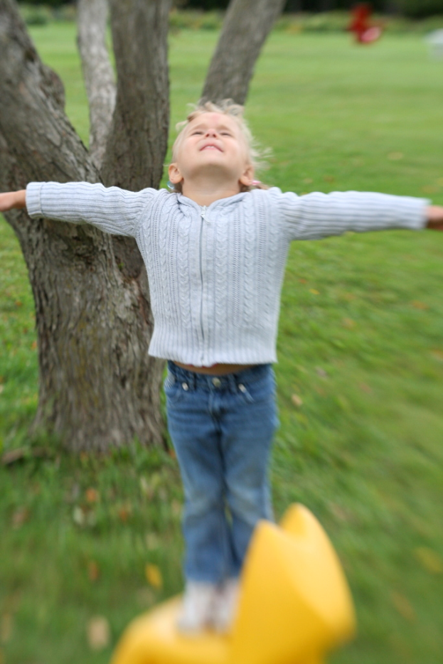 Ein Kind in Jeans streckt beide Hände, während es auf einem gelben Objekt steht, mit einem Baum und Gras im Hintergrund.