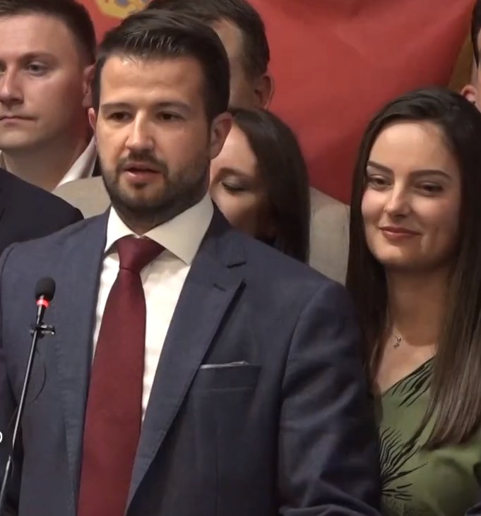 Ein Mann im Anzug und Krawatte spricht an einem Podium mit einem Mikrofon zu einer Menge hinter einem Banner, wahrscheinlich über ein Regierungsverbot von Social Media.