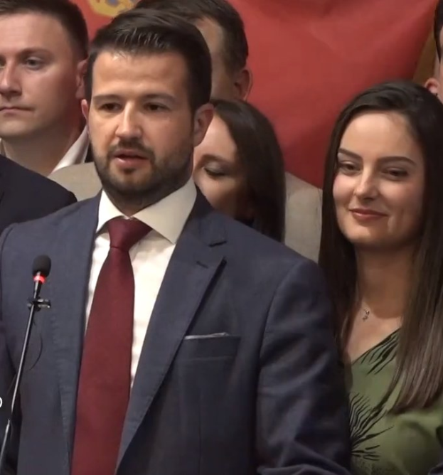 Ein Mann im Anzug und Krawatte spricht an einem Podium mit einem Mikrofon zu einer Menge hinter einem Banner, wahrscheinlich über ein Regierungsverbot von Social Media.