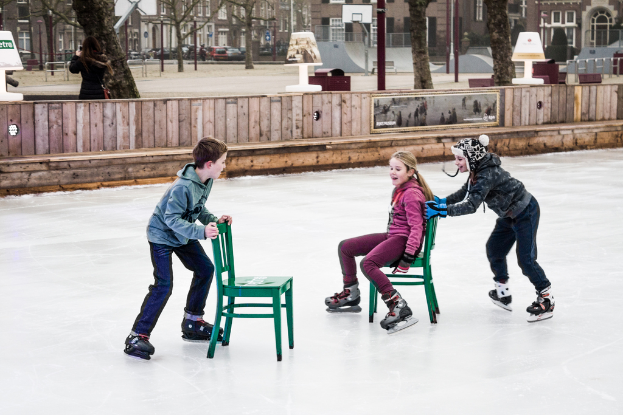 Kinder beim Skifahren im Vordergrund eines Spielplatzes mit drei Kindern und zwei StÃ¼hlen in der Mitte und GebÃ¤uden, BÃ¤umen, BÃ¤nken, PfÃ¤hlen und einem Basketballfeld im Hintergrund.