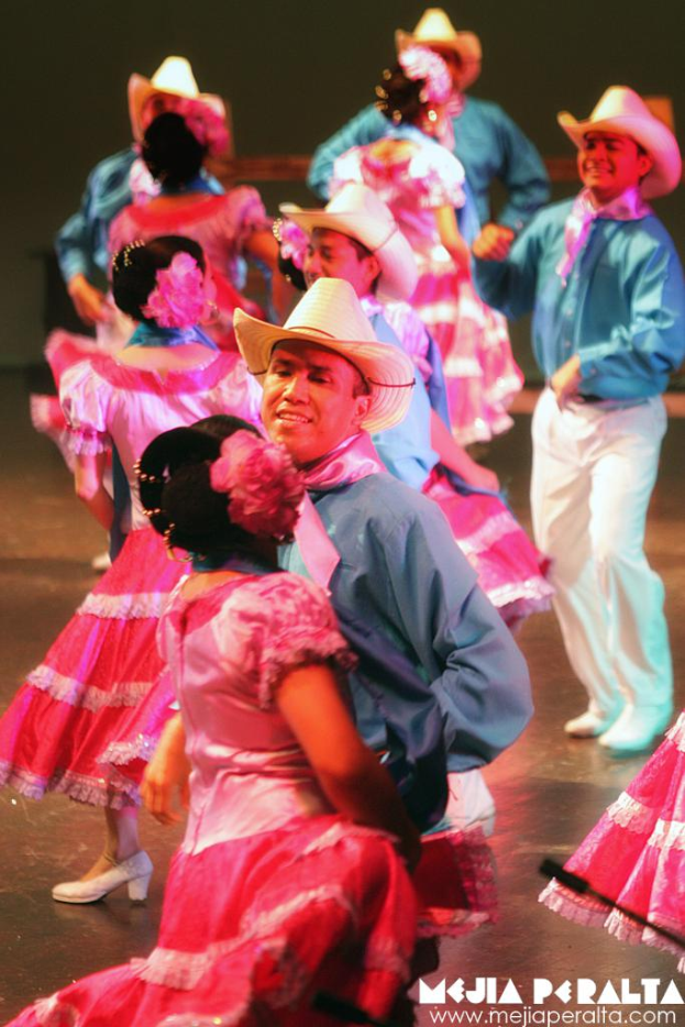 Eine Gruppe von Menschen in blauen Hemden und Hüten tanzt neben einer Gruppe in pinken Kleidern, mit Text in der rechten unteren Ecke.