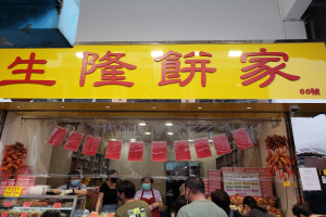 Eine Gruppe von Menschen steht vor einem chinesischen Restaurant mit Schildern auf Chinesisch, einige tragen Masken, mit Essen in Glasvitrinen und Papieren auf der rechten Seite.