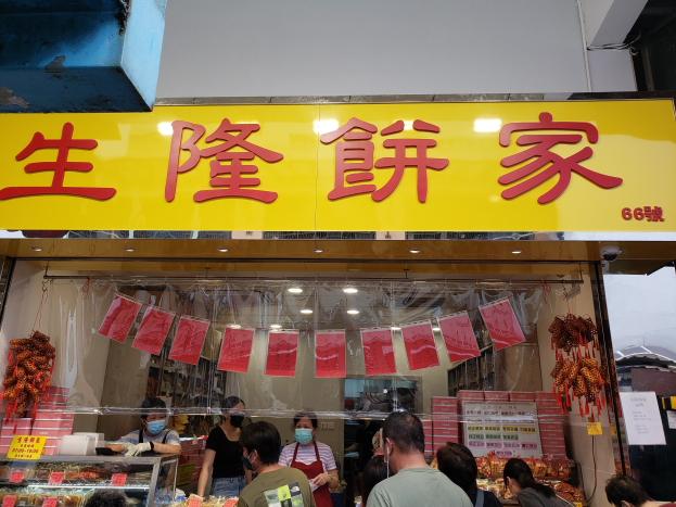 Eine Gruppe von Menschen steht vor einem chinesischen Restaurant mit Schildern auf Chinesisch, einige tragen Masken, mit Essen in Glasvitrinen und Papieren auf der rechten Seite.