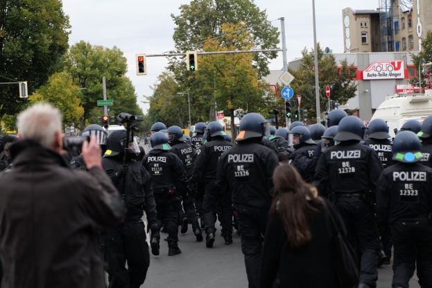 Polizisten in schwarzen Uniformen und Helmen gehen über eine Straße vorbei an einer Menge, einige halten Kameras, mit Bäumen, Gebäuden und einem klaren Himmel im Hintergrund.
