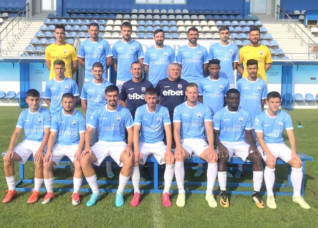 Eine Gruppe von Männern, einige sitzen auf Bänken und andere stehen auf Gras, auf einem Fußballfeld mit Tribünen und Gebäudestrukturen im Hintergrund.