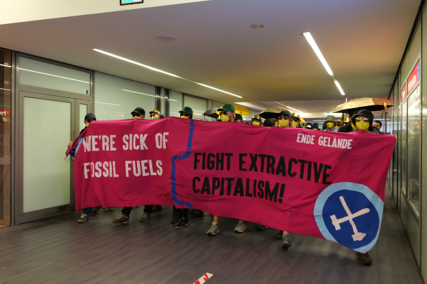 Eine Gruppe maskierter Personen mit einem Banner, auf dem "Wir haben genug von fossilen Brennstoffen, kämpft gegen den extraktiven Kapitalismus" steht, vor einer Wand mit Glastüren und Deckenleuchten.