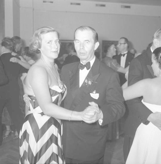 Zwei Menschen, ein Mann in einem schwarzen Blazer und weißem Hemd mit schwarzer Hose und eine Frau in einem unbekannten Outfit, tanzen zusammen auf einer formellen Veranstaltung, halten sich an den Händen und wiegen sich im Takt gegen eine Wand im Hintergrund.