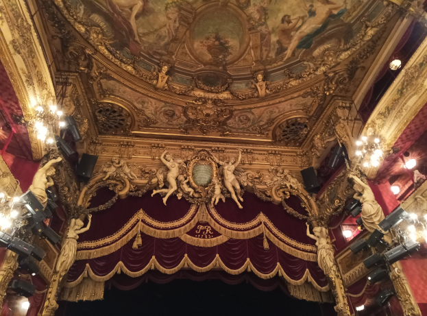 Innere eines Theaters mit einer roten Gardine, Kronleuchtern, Skulpturen, Lichtern und einer Deckenmalerei, die eine Bühne und ein paar Menschen unten zeigt.