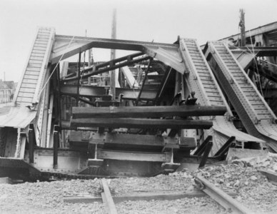 Schwarz-weiß-Foto einer Bahnschiene, die zu einer baufälligen Brücke mit zerbrochenen Geländern und Schutt führt, umgeben von Gebäuden unter einem klaren Himmel.