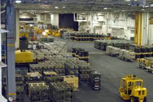 Ein großes Lagerinnere mit zahlreichen Lastwagen, Containern, Menschen, Lampen, Stangen und einem prominenten Rad.