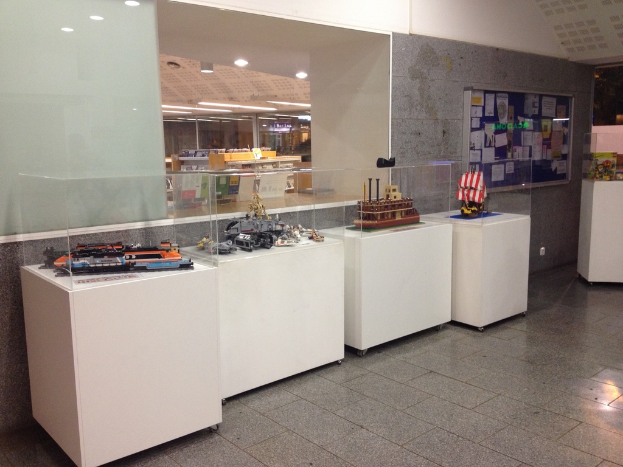 Auf weißen Tischen in einem Ausstellungssaal ausgestellte Gegenstände, mit einer Wandtafel voller Plakate dahinter und einer Decke oben im Bild.