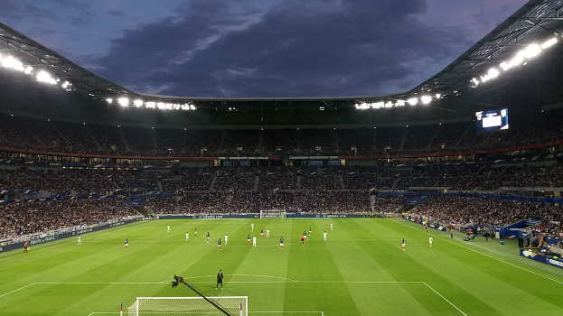 Ein Fußballspiel wird in einem großen Stadion mit Menschen auf dem Boden, einem Tor, einer Kamera mit Stativ, Bannern mit Text, einem Dach mit Deckenleuchten und einem Anzeigebildschirm unter einem bewölkten Himmel gespielt.