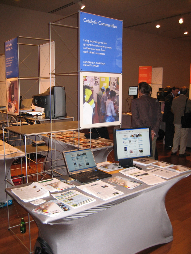 Ein Ladeneinrichtungsbild mit einem zentralen Tisch, auf dem Bücher, ein Laptop und ein Desktop-Computer liegen, umgeben von Bücherständern, mit Werbetafeln, einem Fernseher und Deckenleuchten im Hintergrund und ein paar Menschen auf der rechten Seite.