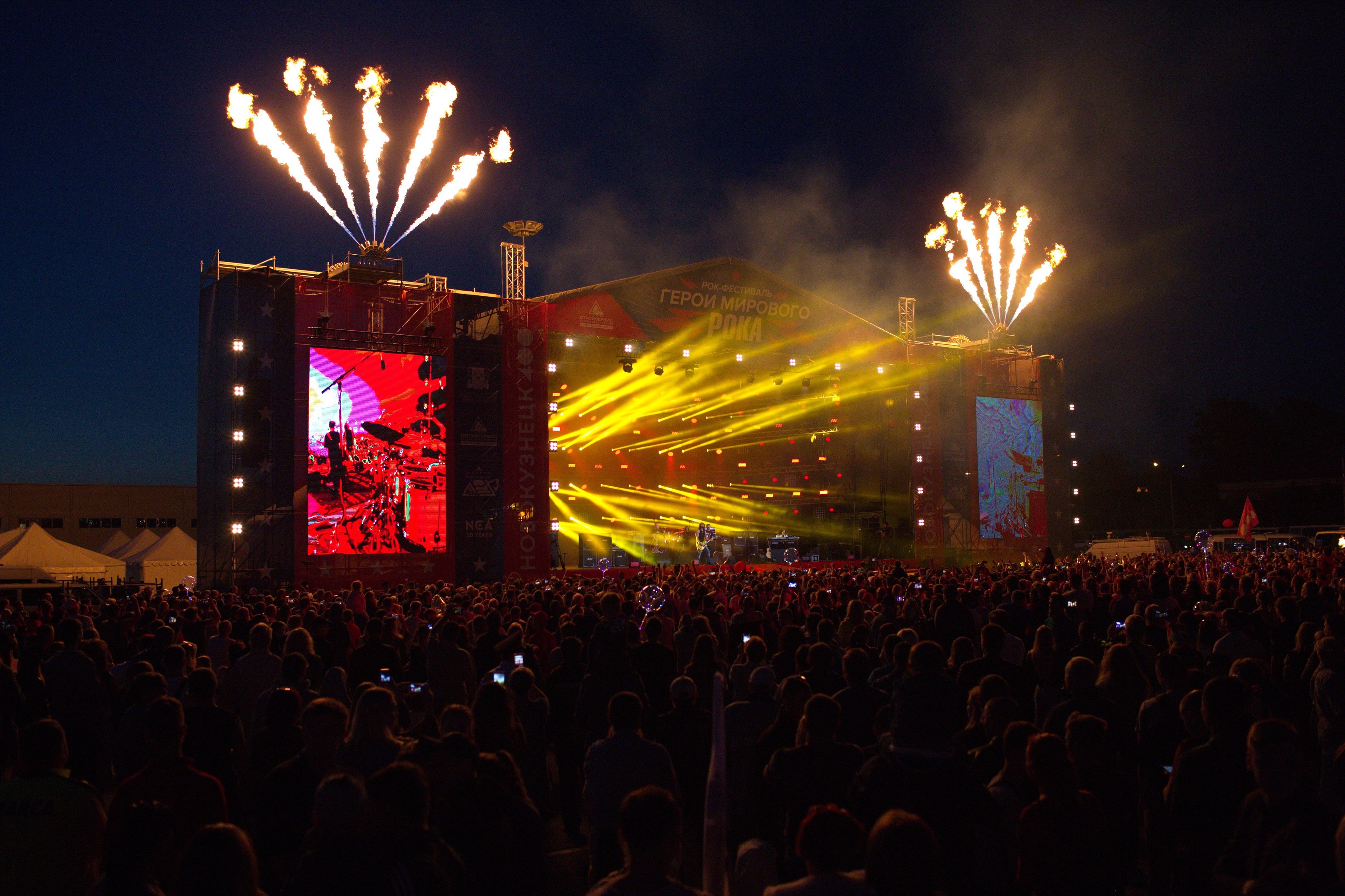 Eine große Menschenmenge auf einem Musikfestival schaut auf einer hell erleuchteten Bühne Performern zu, mit Zelten links und Feuerwerk am dunklen Himmel dahinter.