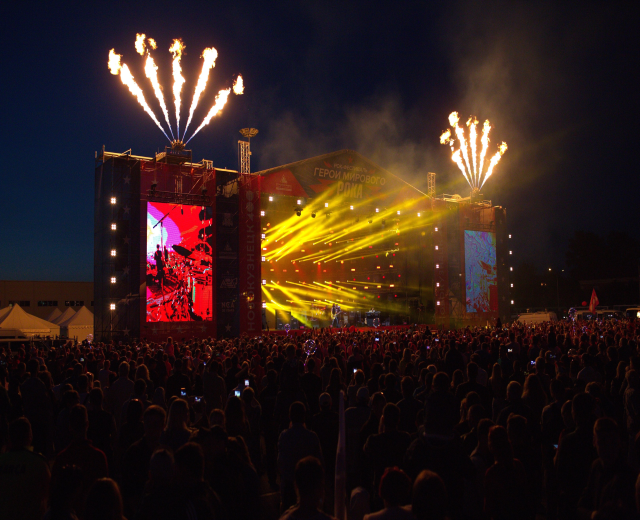 Eine große Menschenmenge auf einem Musikfestival schaut auf einer hell erleuchteten Bühne Performern zu, mit Zelten links und Feuerwerk am dunklen Himmel dahinter.