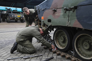 Zwei Männer in Militäruniform arbeiten an einem Fahrzeug in einer Garage, umgeben von Werkzeugen, mit anderen Fahrzeugen und einer Hütte im Hintergrund.