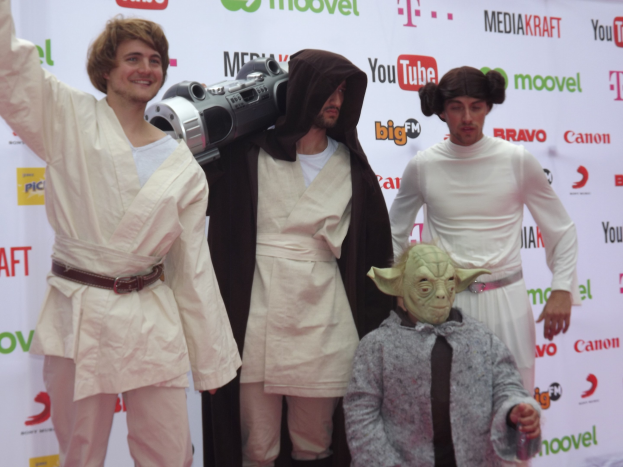 Eine Gruppe von Menschen in Star-Wars-Kostümen, die auf einem roten Teppich posieren, wobei einer ein Objekt in der Mitte hält und ein Banner mit Text und Logos im Hintergrund zu sehen ist.