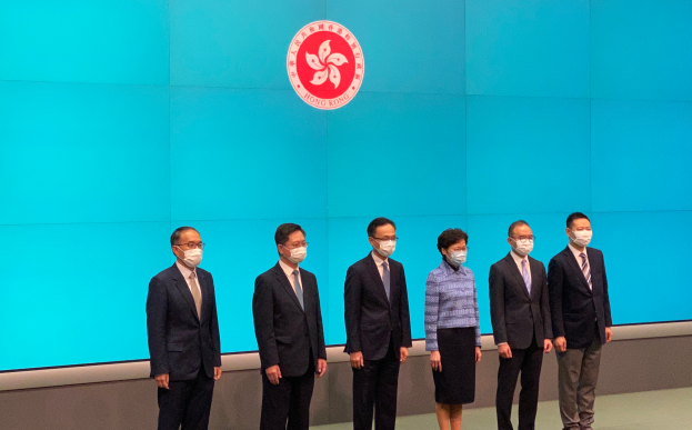 Eine Gruppe von Menschen in Masken steht nebeneinander vor einer blauen Wand mit einem Logo und nimmt an einer Pressekonferenz in Hong Kong teil.